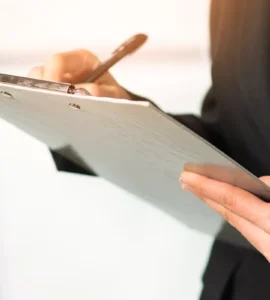 Pessoa com traje social escrevendo em uma prancheta, destacando mãos, caneta e documento em ambiente corporativo.