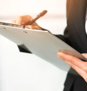 Pessoa com traje social escrevendo em uma prancheta, destacando mãos, caneta e documento em ambiente corporativo.