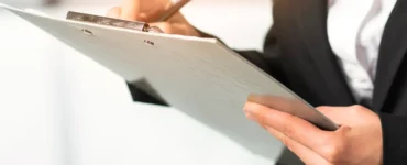 Pessoa com traje social escrevendo em uma prancheta, destacando mãos, caneta e documento em ambiente corporativo.