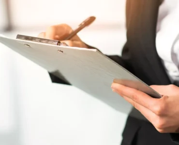 Pessoa com traje social escrevendo em uma prancheta, destacando mãos, caneta e documento em ambiente corporativo.