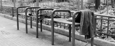 Banco de praça com barras metálicas divisórias impedindo que pessoas se deitem, com roupas deixadas sobre o assento.