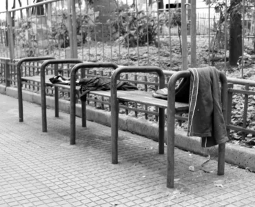 Banco de praça com barras metálicas divisórias impedindo que pessoas se deitem, com roupas deixadas sobre o assento.