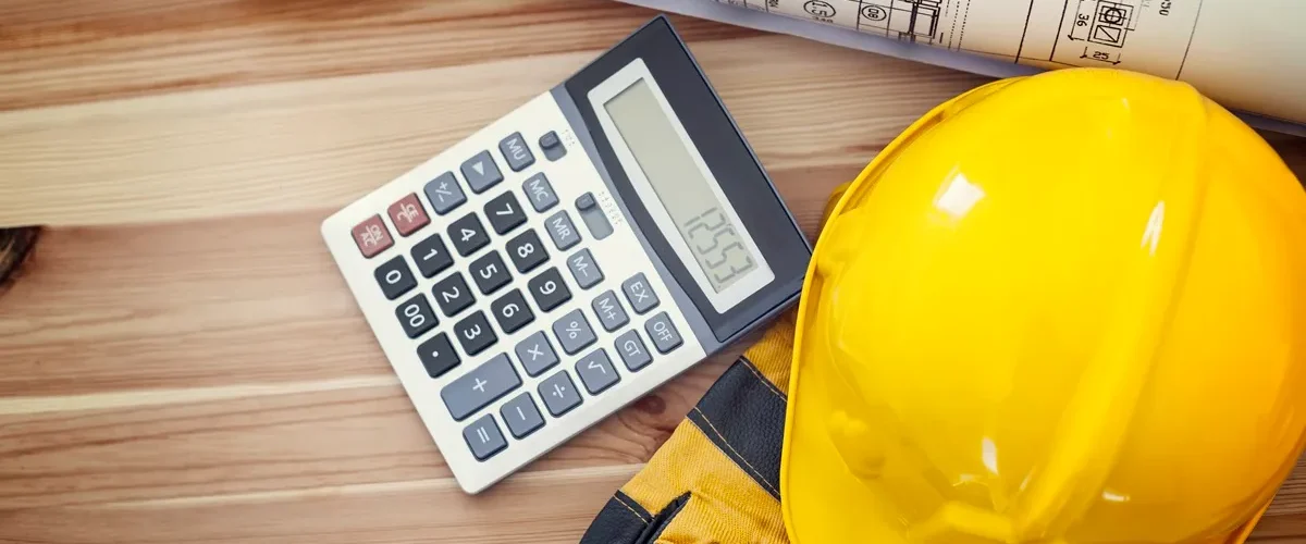 Capacete de segurança amarelo, luvas de proteção, calculadora e planta arquitetônica sobre mesa de madeira, representando orçamento e planejamento de obras.