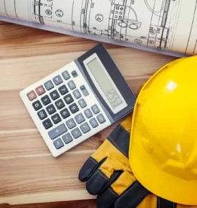 Capacete de segurança amarelo, luvas de proteção, calculadora e planta arquitetônica sobre mesa de madeira, representando orçamento e planejamento de obras.