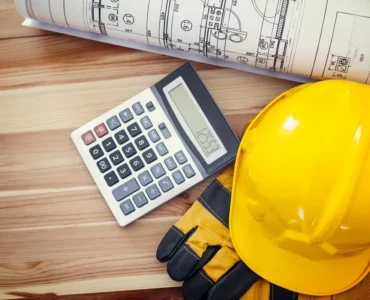 Capacete de segurança amarelo, luvas de proteção, calculadora e planta arquitetônica sobre mesa de madeira, representando orçamento e planejamento de obras.
