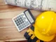 Capacete de segurança amarelo, luvas de proteção, calculadora e planta arquitetônica sobre mesa de madeira, representando orçamento e planejamento de obras.