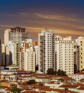 Panorama urbano com prédios altos e bairro de casas térreas ao entardecer, sob um céu alaranjado.