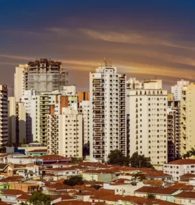 Panorama urbano com prédios altos e bairro de casas térreas ao entardecer, sob um céu alaranjado.