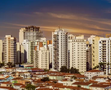 Panorama urbano com prédios altos e bairro de casas térreas ao entardecer, sob um céu alaranjado.