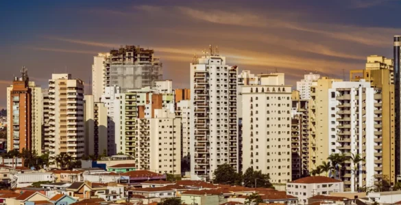 Panorama urbano com prédios altos e bairro de casas térreas ao entardecer, sob um céu alaranjado.