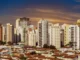 Panorama urbano com prédios altos e bairro de casas térreas ao entardecer, sob um céu alaranjado.