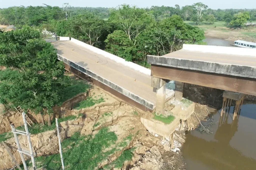 Ponte desabada sobre rio com margens erodidas, causada por seca prolongada e instabilidade do solo