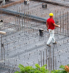 Dois trabalhadores com capacetes caminhando sobre armações de ferro em uma obra de construção civil.