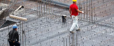 Dois trabalhadores com capacetes caminhando sobre armações de ferro em uma obra de construção civil.