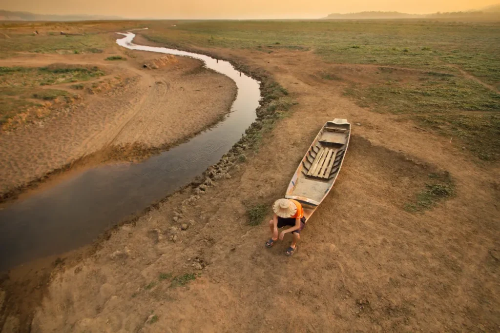 Pessoa sentada ao lado de um barco em área de rio seco com pouca água e solo árido