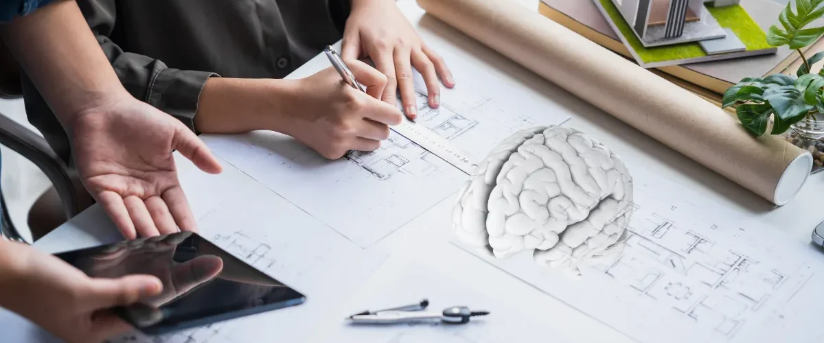 Pessoa desenhando planta arquitetônica sobre mesa com maquete de casa e modelo de cérebro ao lado