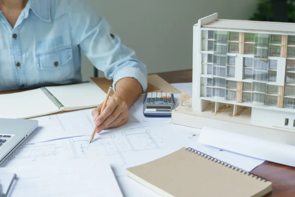 Pessoa desenhando planta de ambiente com maquete de prédio e materiais de trabalho sobre a mesa