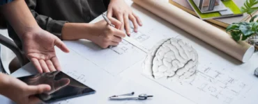 Pessoa desenhando planta arquitetônica sobre mesa com maquete de casa e modelo de cérebro ao lado