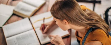 estudante escrevendo em caderno com vários livros abertos sobre a mesa de madeira