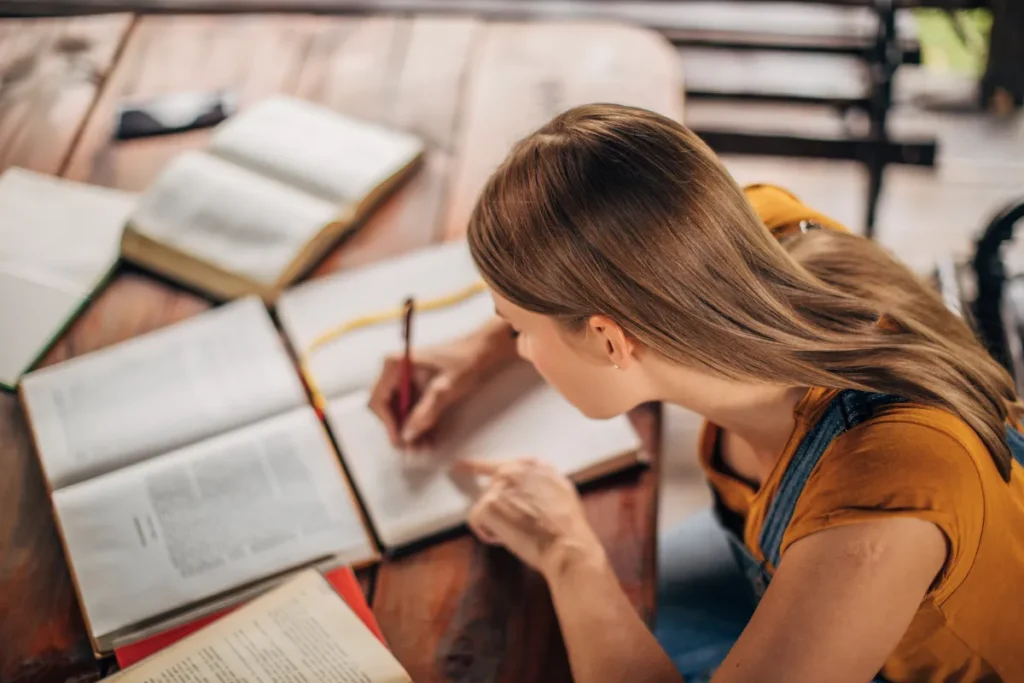 Estudante escrevendo em um caderno enquanto consulta vários livros abertos sobre a mesa.
