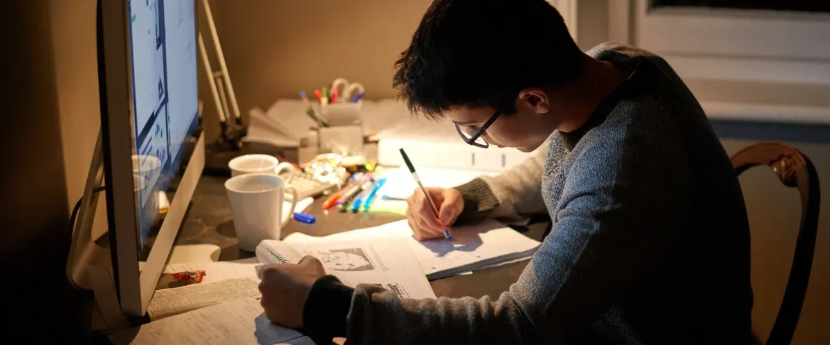 Estudante sentado à mesa estudando e fazendo anotações enquanto consulta materiais no computador.