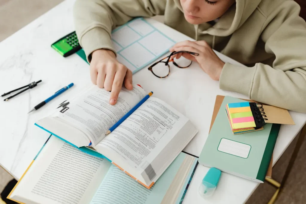 Pessoa apontando para um livro aberto enquanto estuda com cadernos, lápis e marcadores sobre a mesa.