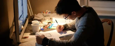 Estudante sentado à mesa estudando e fazendo anotações enquanto consulta materiais no computador.