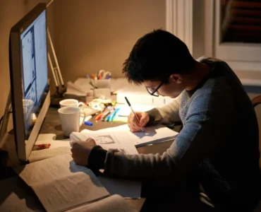Estudante sentado à mesa estudando e fazendo anotações enquanto consulta materiais no computador.