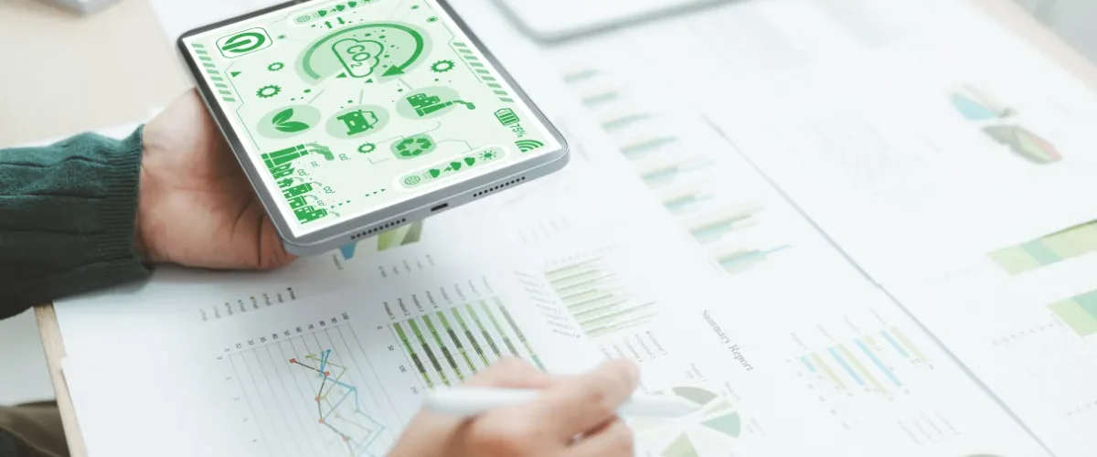 Pessoa segurando um tablet com ícones de sustentabilidade enquanto analisa gráficos e relatórios sobre uma mesa.
