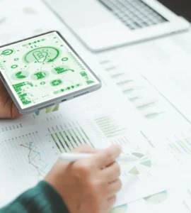 Pessoa segurando um tablet com ícones de sustentabilidade enquanto analisa gráficos e relatórios sobre uma mesa.