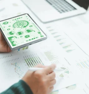 Pessoa segurando um tablet com ícones de sustentabilidade enquanto analisa gráficos e relatórios sobre uma mesa.