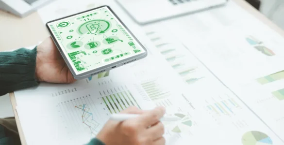 Pessoa segurando um tablet com ícones de sustentabilidade enquanto analisa gráficos e relatórios sobre uma mesa.