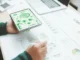 Pessoa segurando um tablet com ícones de sustentabilidade enquanto analisa gráficos e relatórios sobre uma mesa.