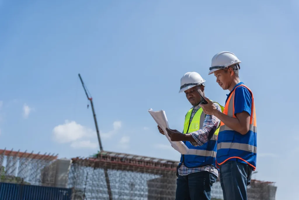 Dois trabalhadores da construção com capacetes e coletes refletivos verificando documentos em um canteiro de obras com estrutura ao fundo