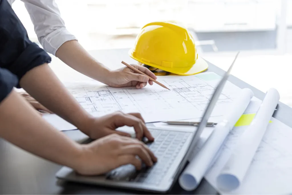 Mãos utilizando um laptop enquanto apontam para uma planta técnica com capacete de segurança ao lado sobre a mesa
