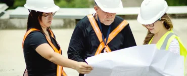 Três profissionais com capacetes e coletes refletivos analisando uma planta de obra em ambiente externo