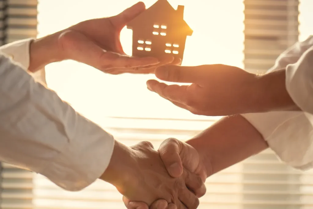 Duas pessoas apertando as mãos enquanto seguram uma miniatura de casa entre elas